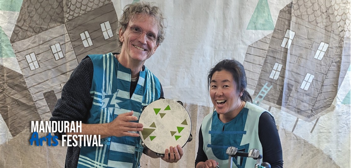 A man and a woman stand in front of a backdrop with cartoon houses painted on it. The man holds a tambourine and they both are smiling broadly